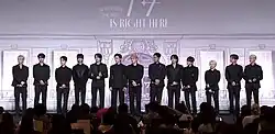 Thirteen men in black suits standing in a line on a stage with press photographers in attendance