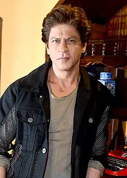 Shah Rukh Khan posing indoors in a mesh-detailed black jacket over a grey shirt.
