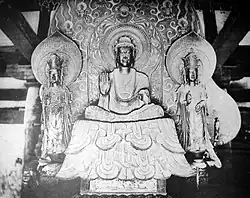 Front view of a central figure sitting cross-legged on a raised platform which is flanked by two smaller standing statues. The central figure has the palm of his right hand turned to the front. The attendants look identical, pointing upwards with their right hand and their left hand lifted halfway, touching the thumb with the middle finger. Each of the three statues has a halo.