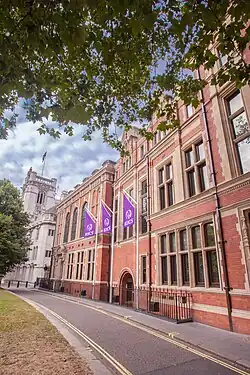 Redbrick building with natural stone highlights and tall stone mullioned windows running away from camera having an exaggerated perspective and proportions with a narrow tarmacced road in front against a dappled sky on a fine day