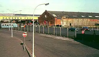 Fenced-off tarmac car parking area, with a run-down red-brick industrial building in the background.