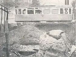 A tram car occupies the top half of the picture. The bottom half shows a several-metre-deep ditch containing soil, boulders, and torn cables or piping.