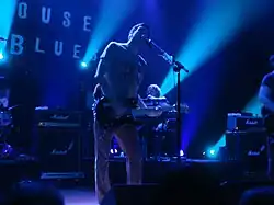 A man playing guitar and singing, with another in the background playing keyboards.