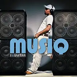 An image of Musiq standing between loudspeakers stacked on top of each other, sporting a white t-shirt, jeans, hat and black headphones.