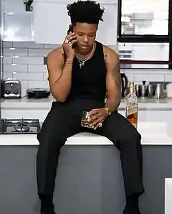 A man in black vest and pants is on a phone call while sitting on a shelf with a half-filled glass of liquor in hand.