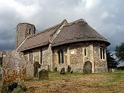 A flint church seen from the southeast, with thatched roofs, an apsidal chancel, a slightly taller nave beyond it, and a tower with an octagonal top