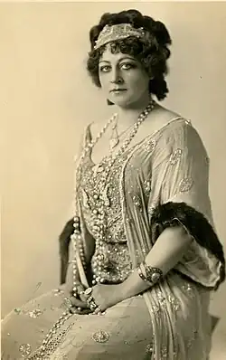 A white woman with curly dark hair piled up loosely and wrapped with a scarf; she is seated and wearing a light-colored gown with extensive beading