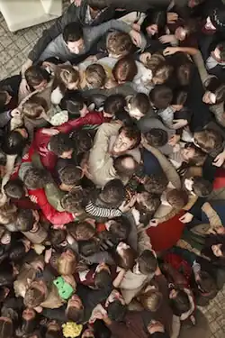 An overhead shot of more than fifty people crowded together, arms out embracing each other in a group hug, their attention on one central figure who is looking upward at the camera.