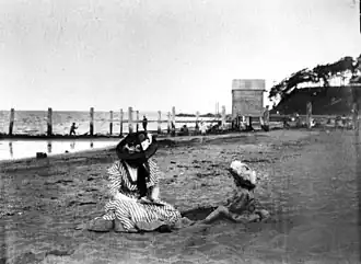 Woman and young child sitting on the beach at Sandgate. The woman appears to be reading to the child, and both are wearing hats. A boatshed and jetty are visible in the background. People are fishing from the jetty.