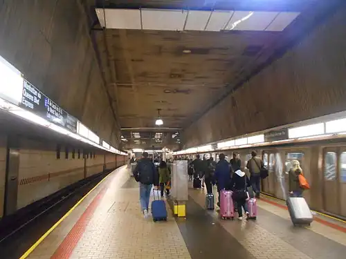 A view of passengers at the upper level, exiting an E train with luggage and heading for the AirTrain to JFK Airport