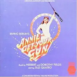 The album cover shows Suzi Quatro with her right hand on her hip and grasping the middle of a rifle in her left hand, raising it up high. She is smiling, wearing a white leather dress plus jacket, and framed by fireworks against a light blue background.