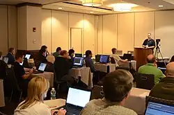 A man speaking from a podium to a room of people with laptop computers