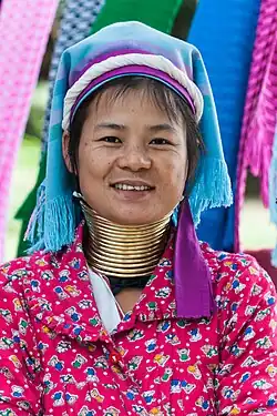 A smiling woman wearing, blue, purple and white along with a matching graphic dress with cartoon bears on it. She has approximately 15 loops of a golden metal stacked around her neck, notably lengthening it. She has a tan skin-tone with dark brown hair. Her hat is made of layered cloth and looks like it was arranged by hand. She appears to have no other body modifications such as ear piercings.