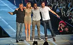 Four people—a middle aged white man in a striped black shirt and jeans, a young white woman in heels and a sparkling dress, a black man in a grey shirt and pants, and a white man in a white tee and jeans, posing together onstage