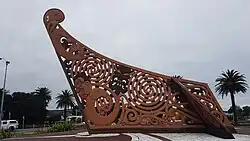 An elaborate Māori carving painted reddish brown carved like the end of a waka canoe set into concrete