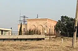 A single-story powerhouse building of the Tetrikhevi HPP in a neoclassical style with arched windows and a pale pink facade. A transmission tower is visible in the background.
