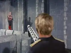 A man in the foreground, operating a machine gun, fires at a man in the background who is wearing a jet pack. Behind the man in the background is the wall of a castle.