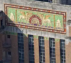 Closeup of a portion of a building, with a multicolored relief decorated with a sunburst, mythological animals, and beehives, on a green background. Below the relief are long piers of windows and brick walls.