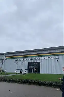 The newly opened High Performance Centre can be seen with some grass in the foreground. The centre resembles a warehouse with the Saints' club colours forming a stripe along the side. In the background, the sky is grey.