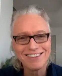 A man with long grey hair tied in a ponytail, wearing black-rimmed glasses and smiling at the camera.