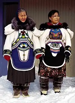 Two Inuit women in clothing covered with detailed beadwork and colourful fabric designs. They have fur ruffs on the hoods of their parkas, cloth skirts beneath their parkas, and fur boots.