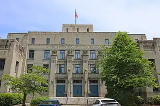 Front of four story concrete building with flag post on roof and two trees in front.