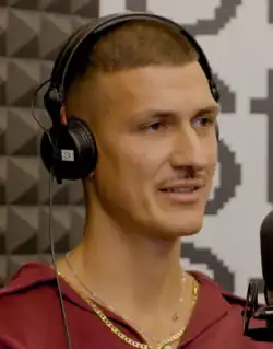 Headshot of Tobias Rahim during an podcast interview. He is a man with short buzz cut and a pencil moustache, wearing a hoodie and a silver and gold necklace.