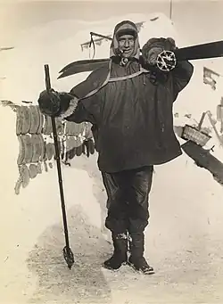 Man, standing, wearing a smock, heavy trousers and boots. He has a ski stick in his right hand, a pair of skis strapped on his back, and is carrying a rounded bundle on his shoulder. Behind him on the ground is assorted polar equipment.