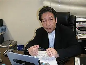 Portrait photo of a Japanese man with short black hair wearing a black business jacket sitting at an office desk.