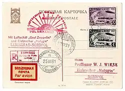 The reverse side of a postcard. A Cyrillic inscription is printed at the top, then "Par Avion". The two stamps are for 30 kopecks (magenta on white) and 1 rouble (black on white). They show a dramatic depiction of the airship above the icebreaker, with a polar bear on a spur of ice in the foreground. At the top is the hammer and sickle of the Soviet Union and the words "Pole du Nord 1931". At the bottom is a legend in Cyrillic script. There are four postmarks; bottom-left says "25 VII 31 Leningrad" and the bottom right "27.8.31 Lorch". An address in Lorch, Germany, is type-written below. At left are three cachets and a "Par Avion" sticker with Cyrillic translation.