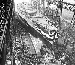 A large warship, still missing most of its superstructure, sits in a dry dock, awaiting its launch. The ship is draped in a large banner and surrounded by crowds of spectators; a huge gantry towers over the ship.