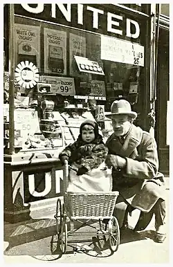 A cigar store with a glass window in the front with cigar packs on display, "UNITED" is written on the glass. In front of the store are an adult man with a baby in a stroller