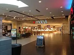 A one-story shopping mall concourse with brown tile flooring. Present is the entrance to a Macy's department store, with signage reading "Macy's"
