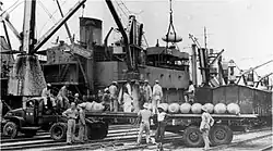 Black and white photograph of a port. Cranes are seen unloading cylindrical structures onto a trailer manned my multiple men, the trailer is parked diagonally on rail lines