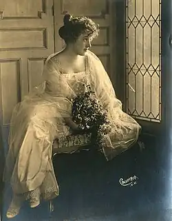 A young white woman wearing a long light dress, sitting at a window
