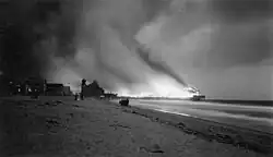 Burning pier and billowing smoke in black and white photograph taken around dusk