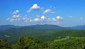 A range of mountains, mostly rounded and covered with green woods, seen past fields in the distance. Above them is a blue sky filled with little fluffy clouds.
