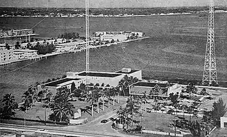 Aerial photograph of two buildings on a man-made island. The larger building on the left has signage saying "WCKT 7 NBC" and WCKT Television" on the sides, while the adjacent building has signage reading "WCKR RADIO". Parking lots are visible in front of the two buildings with trees lining the shared entrance and the street in the foreground. In front of the building on the left and behind the building on the right are two radio towers; the tower on the left is a guyed tower with supporting wires on all sides, while the tower on the right is wider and self-supporting.