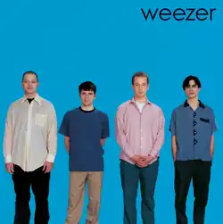 Four plainly dressed young white men stand facing the viewer against a solid blue background, with the band name written in black in the top right corner.