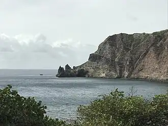 A dark blue bay beyond some bushes on a cloudy day. On the opposite side of the bay is a very large cliff, which curves downside to the left into some pointy rock formations.