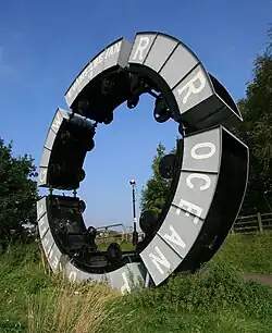 Modern sculpture using wheeled tubs from mineral mining attached to each other forming a raised circle against a bright blue sky