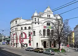 A large building, partly obscured by trees. At its front are two circular towers with turrets, flanking a high pediment. The side visible to the left presents archways and windows.