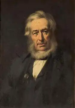Head and torso portrait of man with white hair and beard wearing a dark suit