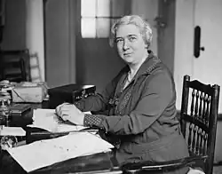 Monochrome photograph of Cullis sitting at her desk and looking over her left shoulder to the camera. There is a parcel and suitcase on the desk.
