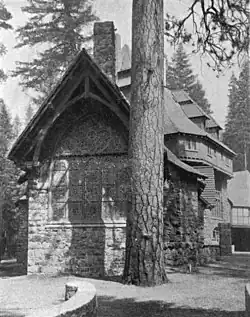 A monochrome photograph of the exterior of a building made of river rock, showing tall stained glass windows at one end, next to the massive trunk of a tall pine tree.
