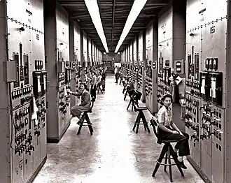 A long corridor with many consoles with dials and switches, attended by women seated on high stools