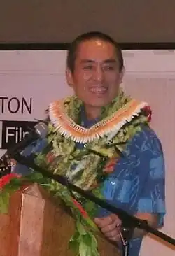 A middle-aged Chinese man standing at a podium, wearing a Hawaiian shirt and lei