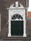 Herplaatste gebeeldhouwde natuurstenen poortomlijsting bekroond door gebroken fronton met Vlissings wapen. Cartouches met emblemen en lettertekens, guirlandes en kop van een moor
