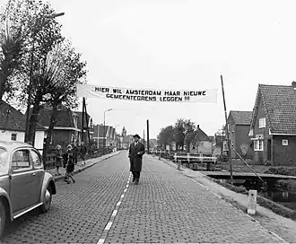 Burgemeester van Landsmeer in 1958 onder spandoek met tekst: 'Hier wil Amsterdam haar nieuwe gemeentegrens leggen'. Sinds 1966 is hier een paar meter vandaan de huidige gemeentegrens van Kadoelen (Gemeente Amsterdam) en Gemeente Landsmeer.