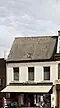 Eenvoudig huis, beneden geheel verbouwd voor winkel, gepleisterde lijstgevel, dwars zadeldak en zijtopgevels met vlechtingen